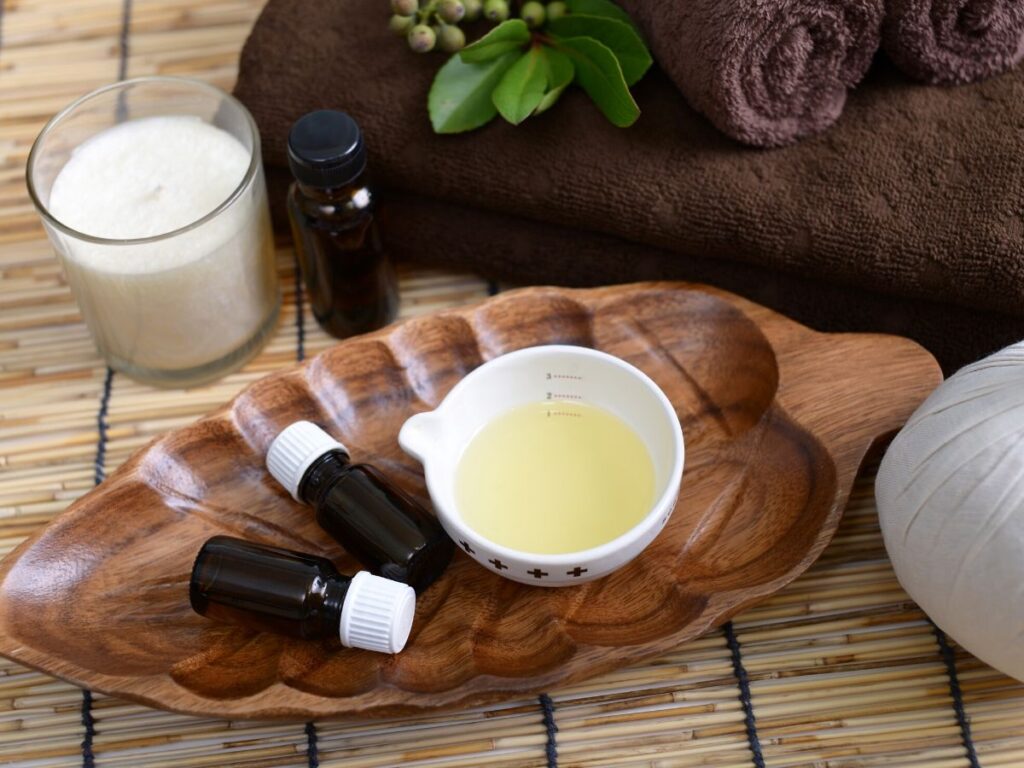 essential oil bottles with carrier oil in a bowl on a wooden leaf platter with candles and towels in background